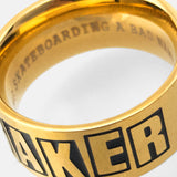 Close-up of a gold Baker Brand Logo Ring with 'BAKER' in black on the outside and 'SKATEBOARDING A BAD' inside.